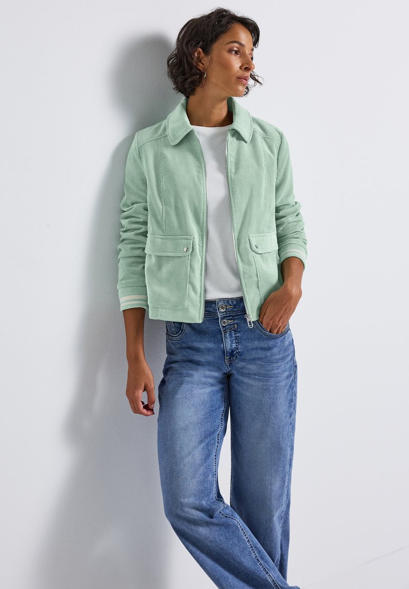 Hellgrüne Cordjacke mit Kragen, zwei Fronttaschen und Reißverschluss, getragen über einem weißen Hemd und kombiniert mit blauen Jeans.