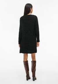 Kvinna med långt mörkt hår, iklädd en strukturerad svart knälång klänning och bruna högklackade stövlar, står vänd bort från kameran på vit bakgrund.