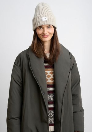 Kobieta w beżowej dzianinowej czapce, ciemnym, oversize'owym płaszczu i swetrze z wzorem, uśmiechająca się na tle jednolitego, jasnego tła.