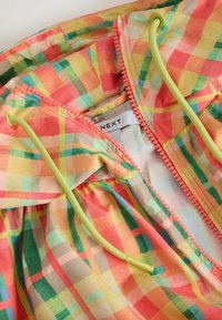 Veste à carreaux colorée avec fermeture éclair orange, cordons verts néon et étiquette de marque "NEXT" visible à l'intérieur du col.