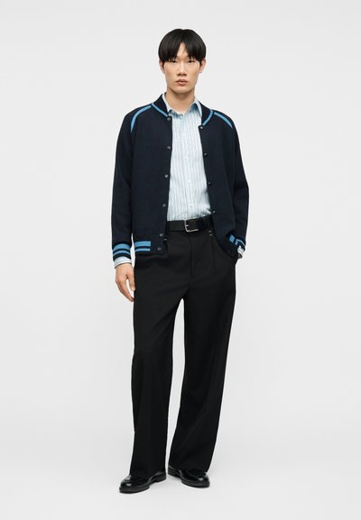Άνδρας που φοράει μία μπλε ναυτική ζακέτα με κουμπιά και ανοιχτό μπλε ριγέ πουκάμισο, μαύρο παντελόνι και μαύρα παπούτσια, στέκεται με το ένα χέρι στην τσέπη.