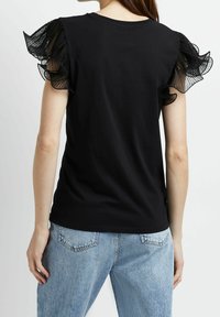 Černé tričko s krátkými, volánkovými průhlednými rukávy. Hladké bavlněné tělo, kulatý výstřih a volný střih, zkombinované se světle modrými džíny.