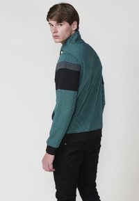 Chaqueta verde y negra con cremallera, fabricada con un tejido texturizado, que presenta bloques de color horizontales en gris y negro. Ajuste slim, puños y dobladillo acanalados.