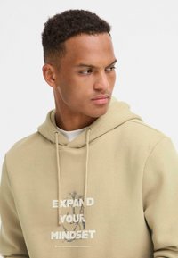 Sudadera beige con capucha ajustable, que presenta el texto "EXPANDE TU MENTALIDAD" en la parte frontal. Material de algodón suave con un corte relajado.