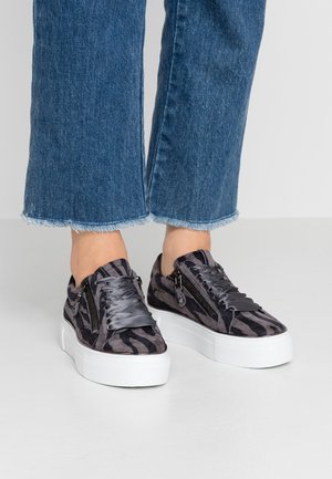Voeten met zwart-grijs gestreepte platformsneakers met dikke witte zolen en ritssluitingdetails, gecombineerd met blauwe spijkerbroeken met rafelige zomen.