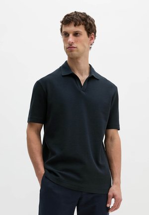 Jeune homme aux cheveux courts et bouclés portant un polo noir à manches courtes, debout, une main dans la poche, regardant sur le côté devant un fond uni.