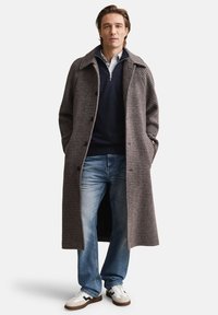 Casaco longo com padrão pied-de-poule castanho, com grandes lapelas, fecho de botões e bolsos laterais, combinado com um suéter azul-marinho, jeans azuis e ténis brancos.