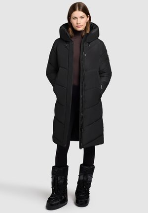 Langer schwarzer Daunenmantel mit Kapuze, mit einem Reißverschluss, seitlichen Taschen und einem gesteppten Design. Trägt schwarze isolierte Stiefel.