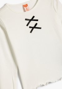 Haut à manches longues blanc à côtes avec un col rond et deux accents en ruban noir formant une croix sur le devant. Les bords sont légèrement effilochés.