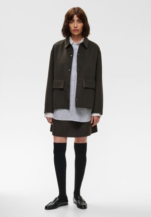 Femme aux cheveux châtain mi-longs portant une veste sombre boutonnée, une chemise rayée, une jupe sombre, des chaussettes noires montantes jusqu’aux genoux et des mocassins noirs, debout sur un fond blanc.