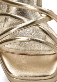 Sandalia de tacón alto metálica dorada con correas entrelazadas y plataforma. Textura suave con un acabado brillante. Cierre no visible.