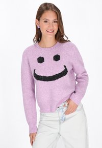 Lila gebreide trui met een zwart smiley gezichtsontwerp, voorzien van een ronde halslijn en lange mouwen, gecombineerd met licht denim jeans.