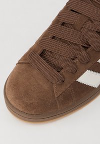 Braune Wildleder-Sneaker mit strukturierter Oberfläche, ausgestattet mit braunen geflochtenen Schnürsenkeln, einer Sohle aus Gummi und weißen Seitenstreifen. Rund gestaltete Zehenpartie.