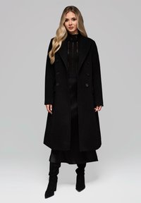 Donna con lunghi capelli biondi che indossa un cappotto nero doppiopetto, una maglietta nera trasparente, una gonna midi e stivali neri con tacco su sfondo grigio.