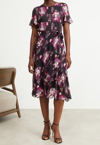 Kvinna som bär en knälång, kortärmad svart klänning med lila och vita abstrakta blommotiv och svarta högklackade skor med ankelsnodd.