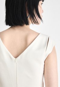 V-neck, sleeveless dress in light cream fabric with smooth texture. Features a back zipper closure and structured shoulder design.