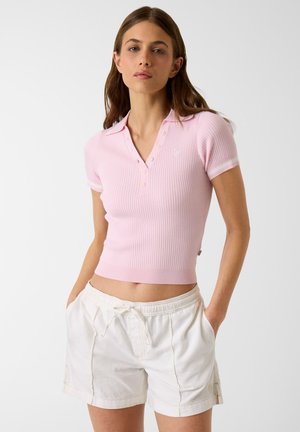 Junge Frau steht mit den Händen in den Taschen, trägt ein eng anliegendes, rosa, geripptes Polo-Shirt mit kurzen Ärmeln und weiße Shorts mit Kordelzug, neutraler Hintergrund.