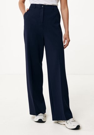 Person trägt marineblaue Hose mit weitem Bein und weiße Sneakers, mit einer Hand in der Tasche und einem weißen Top, das an der Taille sichtbar ist.