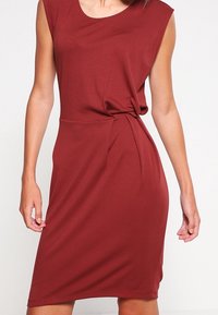 Robe bordeaux en tissu léger avec un col rond, des manches courtes et un détail de nœud sur le côté à la taille. Texture lisse.