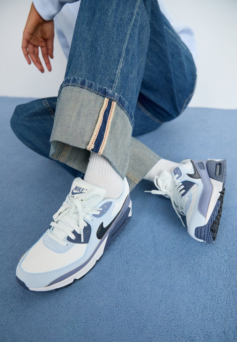 Le sneakers Nike Air Max presentano un design bianco, azzurro chiaro e grigio scuro, realizzato in materiali in mesh e sintetici. Logo swoosh nero sui lati.