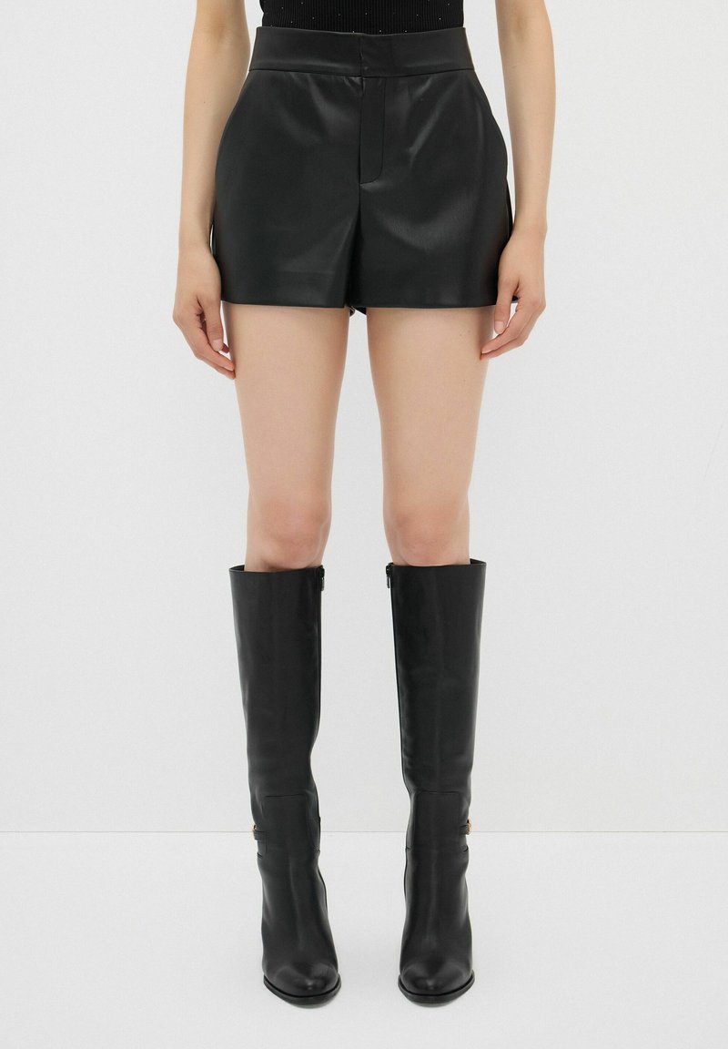 Pantaloni corti di pelle nera con vita alta, texture liscia e vestibilità su misura, abbinati a stivali neri al ginocchio dal design elegante.