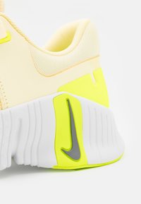 Achteraanzicht van een geel-witte Nike sportschoen met de gestructureerde zool en een grijze Nike-swoosh op een neongele accenten.
