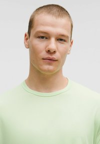 Mladý muž s krátkymi svetlohnedými vlasmi, oblečený v svetlozelenej tričku s okrúhlym výstrihom, hľadí priamo dopredu s neutrálnym výrazom, na jednoduchom svetlošedom pozadí.