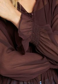 Blusa marrón transparente con mangas abullonadas, cierre de botones y detalle de cintura fruncida. La tela tiene una textura suave y ligera.