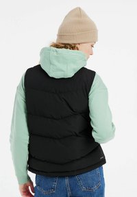 Juodas pūkinis liemenė su siūtomis panelėmis, uždėta ant šviesiai žalio hoodie. Modelis dėvi smėlio spalvos kepurę. Džinsiniai džinsai matomi ties liemeniu.