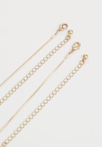 Divi zeltaini ķēžu rokassprādzes ar omāra sprādzi un regulējamām kārtīgām paplašināšanas ķēdēm uz baltā fona.