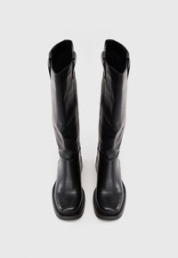 Par de botas altas de couro preto com bicos quadrados e alças, em pé sobre um fundo branco liso.