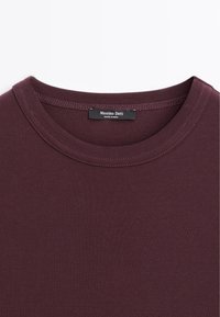 T-shirt bordeaux con collo a giro ed evidenti cuciture, con un'etichetta nera che riporta la scritta "Massimo Dutti Made in India" all'interno del colletto.