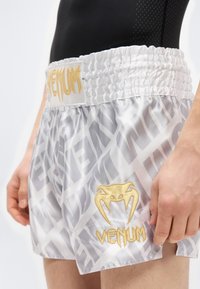 Λευκό σατέν σορτς πυγμαχίας με γκρι επαναλαμβανόμενο μοτίβο και χρυστό κεντημένο λογότυπο «Venum» στη μέση και το τελείωμα.