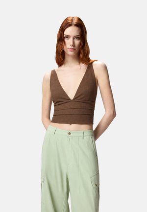 Donna con capelli rossi che indossa un top corto marrone senza maniche con scollo a V profondo e pantaloni larghi verde chiaro con tasche.