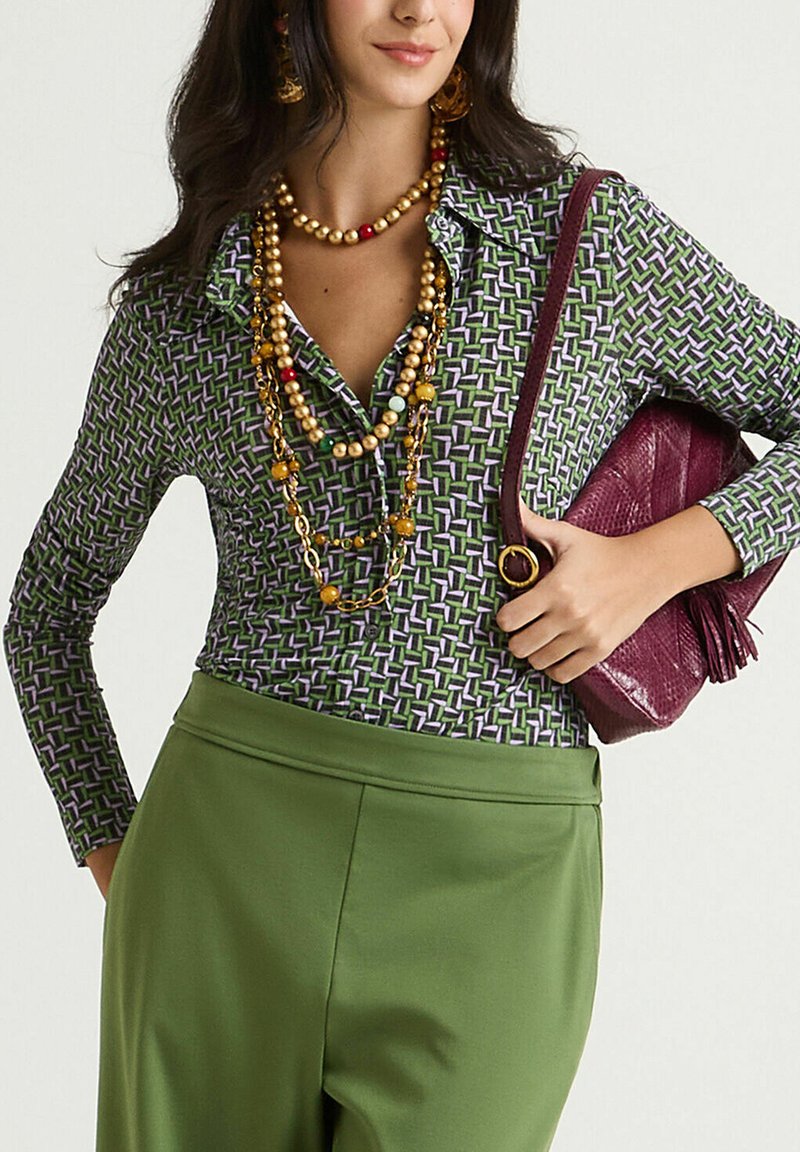Camicia a maniche lunghe con motivo geometrico in verde e viola, abbinata a pantaloni verdi a vita alta. Gli accessori comprendono diverse collane di perline dorate e una borsa bordeaux con dettagli a frangia.