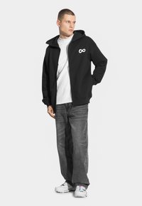 Czarna bluza z kapturem zapinana na zamek, z białym logo nieskończoności, noszona na białej koszulce, zestawiona z luźnymi czarnymi dżinsami i białymi sportowymi butami.