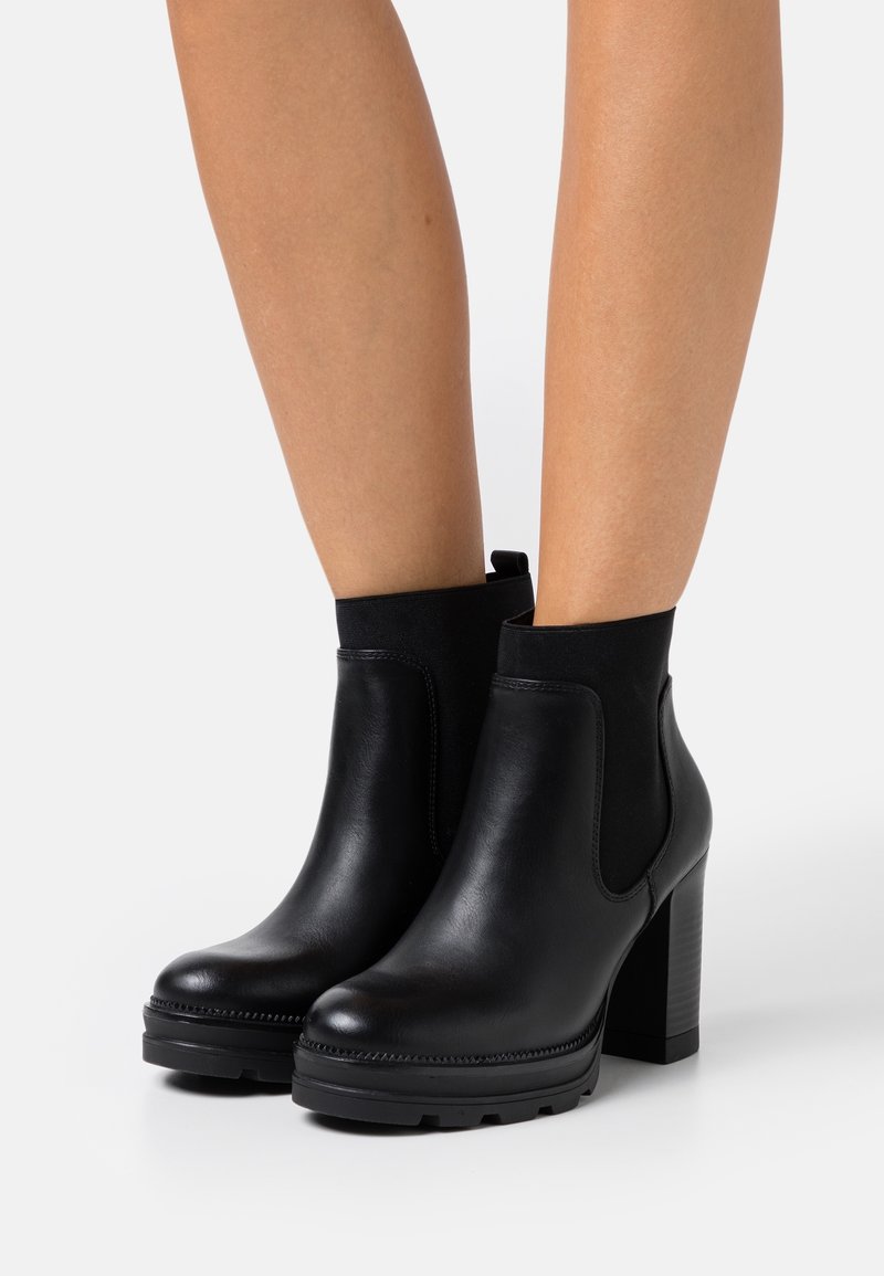 Jambes portant des bottines en cuir noir avec des talons hauts épais et des empiècements latéraux élastiques sur fond blanc.