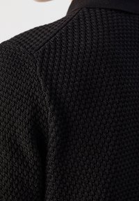 Gros plan sur un tissu tricoté noir texturé sur l'épaule et le haut du dos d'un vêtement, avec une couture visible et un motif gaufré.