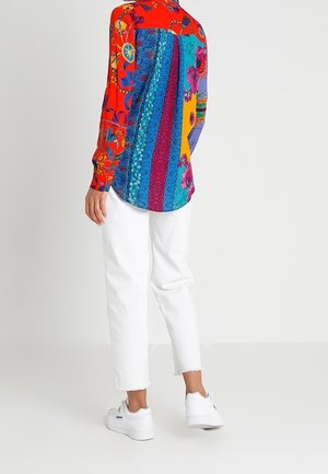 Osoba ubrana w wielokolorową, wzorzystą koszulę z długim rękawem, białe krótkie spodnie i białe trampki, stojąca na tle jednolitego, jasnego tła.