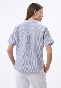 Camicia a maniche corte a righe blu e bianche, realizzata in tessuto leggero, con una piega sul retro e un orlo curvo.