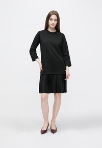 Robe noire avec un col rond et des manches trois-quarts, dotée d'une jupe plissée. Le modèle porte des talons bordeaux.