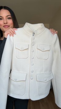 Ženska drži belo teksturirano srajco z gumbi, ki ima štiri sprednje žepne plošče, in stoji v notranjosti ob enostavni steni.