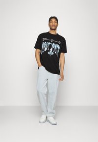 Camiseta negra con texto gráfico azul, combinada con jeans de mezclilla claros y zapatillas blancas. Diseño simple, ajuste casual, mangas cortas.