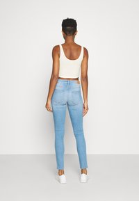 Top corto de color beige claro con tirantes anchos, combinado con unos vaqueros ajustados de color azul claro. Los vaqueros tienen bolsillos traseros y una etiqueta de marca de cuero marrón.