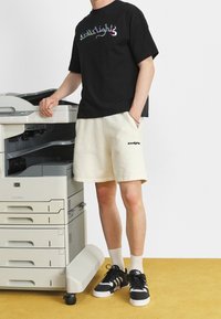 Person in einem schwarzen Grafik-T-Shirt und beigefarbenen Shorts steht neben einem HP Multifunktionsdrucker auf einem gelben Boden vor einer weißen Wand.
