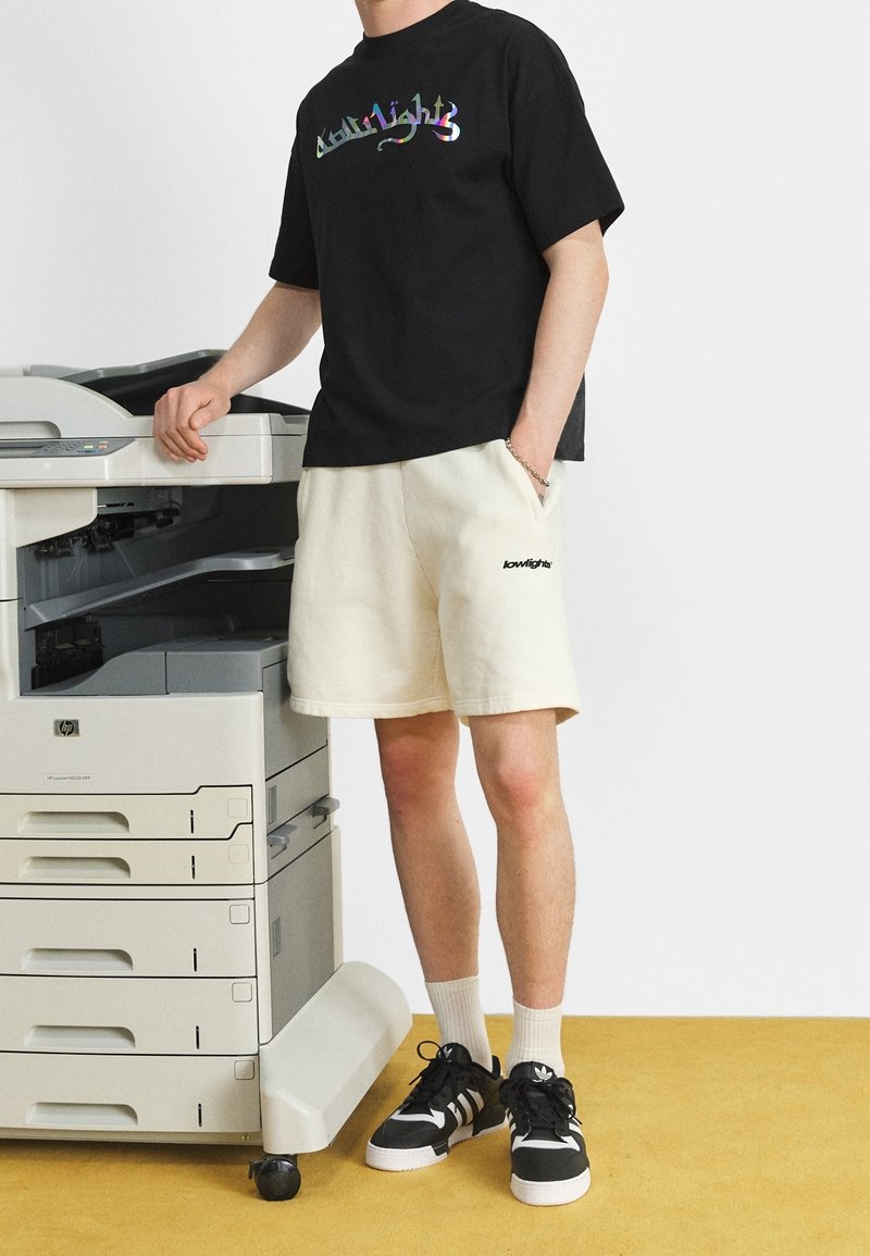 Person in einem schwarzen Grafik-T-Shirt und beigefarbenen Shorts steht neben einem HP Multifunktionsdrucker auf einem gelben Boden vor einer weißen Wand.
