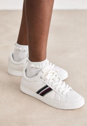 Weiße Low-Top-Sneakers mit roten, schwarzen und weißen Streifendetails, getragen mit weißen, gerafften Knöchelsocken auf brauner Haut.