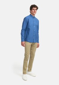 Camisa azul de botones con cuello, mangas largas y un bolsillo, combinada con pantalones caqui y zapatillas blancas. Fondo neutro.