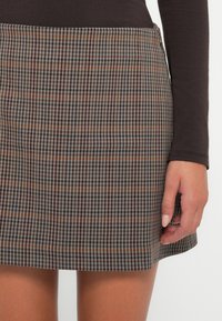 Rutet mini skjørt i grå, brun og oransje toner. Glatt tekstur, tettsittende design med midje, og avsluttet kant. En hånd er delvis synlig.