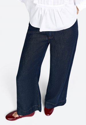 Pantaloni in denim a gamba larga di color blu scuro con una trama sottile. L'orlo presenta una finitura pulita. Abbinati a ballerine rosse lucide.